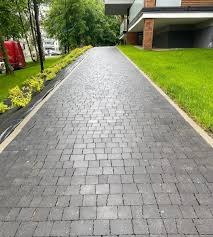 Długa, szara ścieżka ułożona z kostki granitowej prowadząca w kierunku nowoczesnego budynku z balkonem, otoczona zielenią i niskimi krzewami.