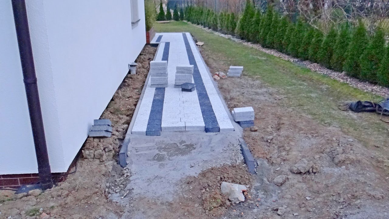 Ścieżka z jasnej kostki brukowej z ciemnymi pasami w trakcie układania, widoczne stosy kostki i przygotowane podłoże, obok domu i trawnika z tujami.
