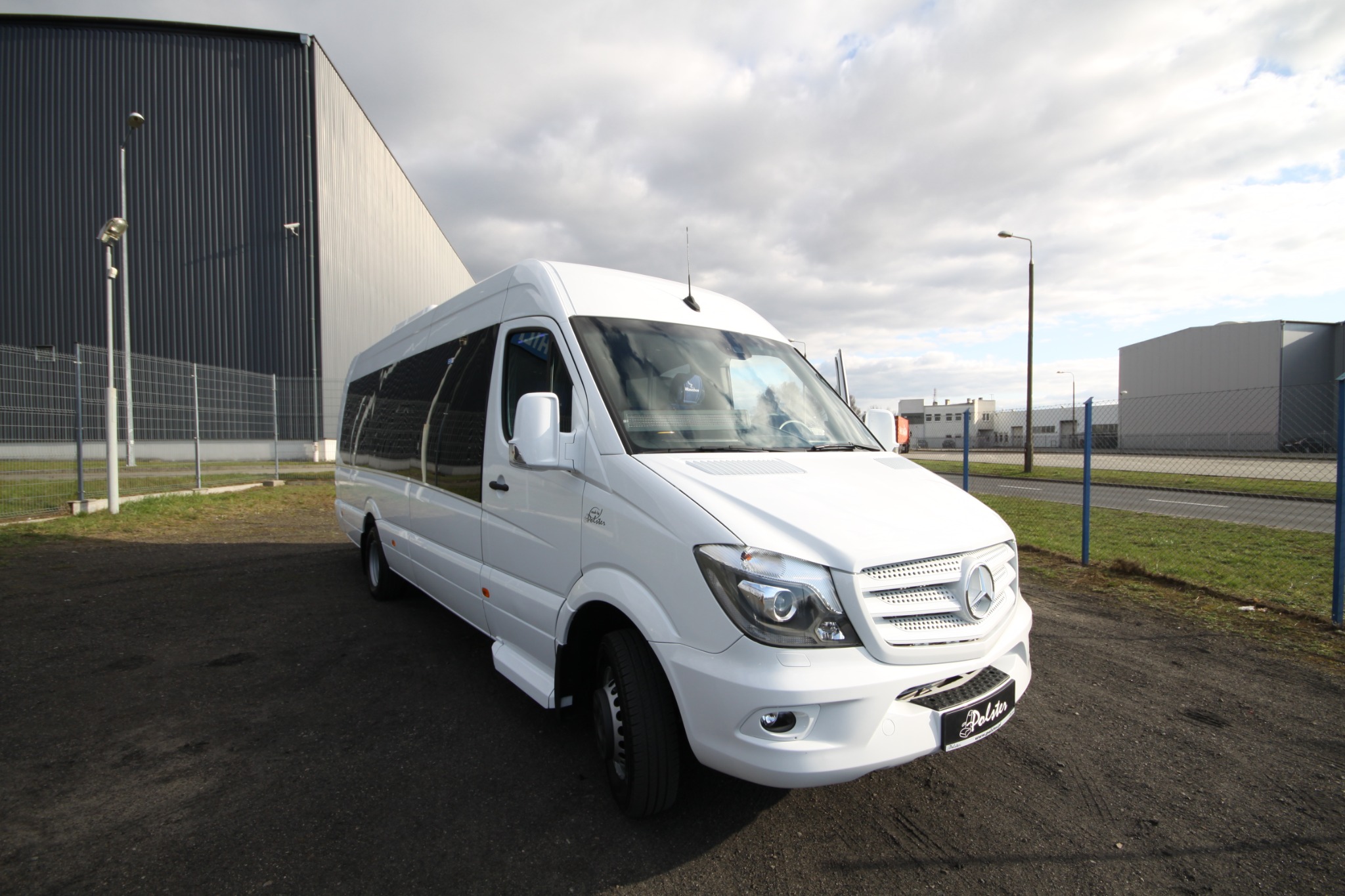 Biały, podwyższony bus Mercedes-Benz Sprinter z przyciemnianymi szybami, zaparkowany na placu przed halą przemysłową w pochmurny dzień.