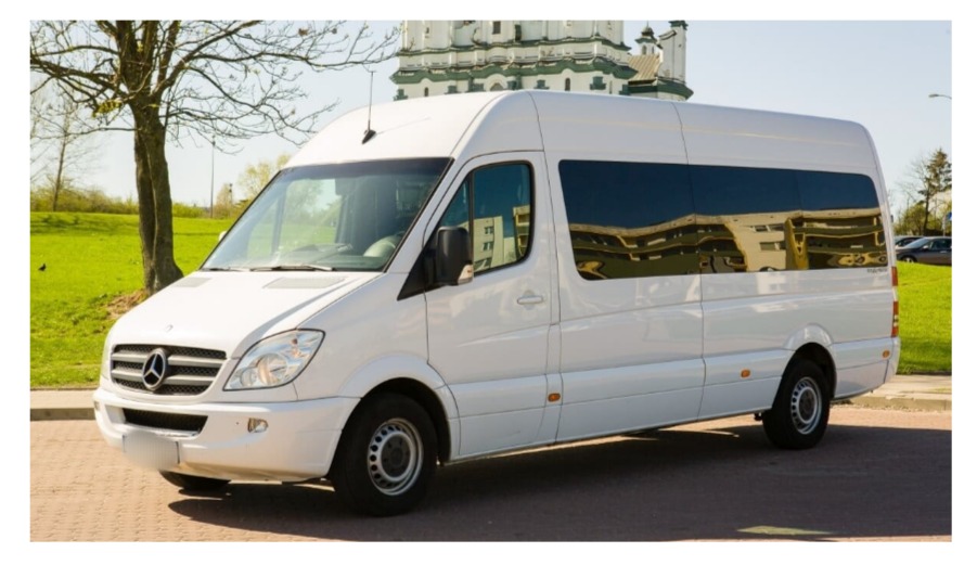 Biały, wysoki bus Mercedes-Benz Sprinter z przyciemnianymi szybami, zaparkowany na brukowanej powierzchni, z budynkiem w tle odbijającym się w szybach.