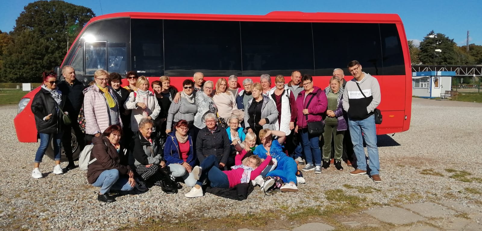 Grupa uśmiechniętych osób pozuje przed czerwonym busem na żwirowym parkingu w słoneczny dzień.