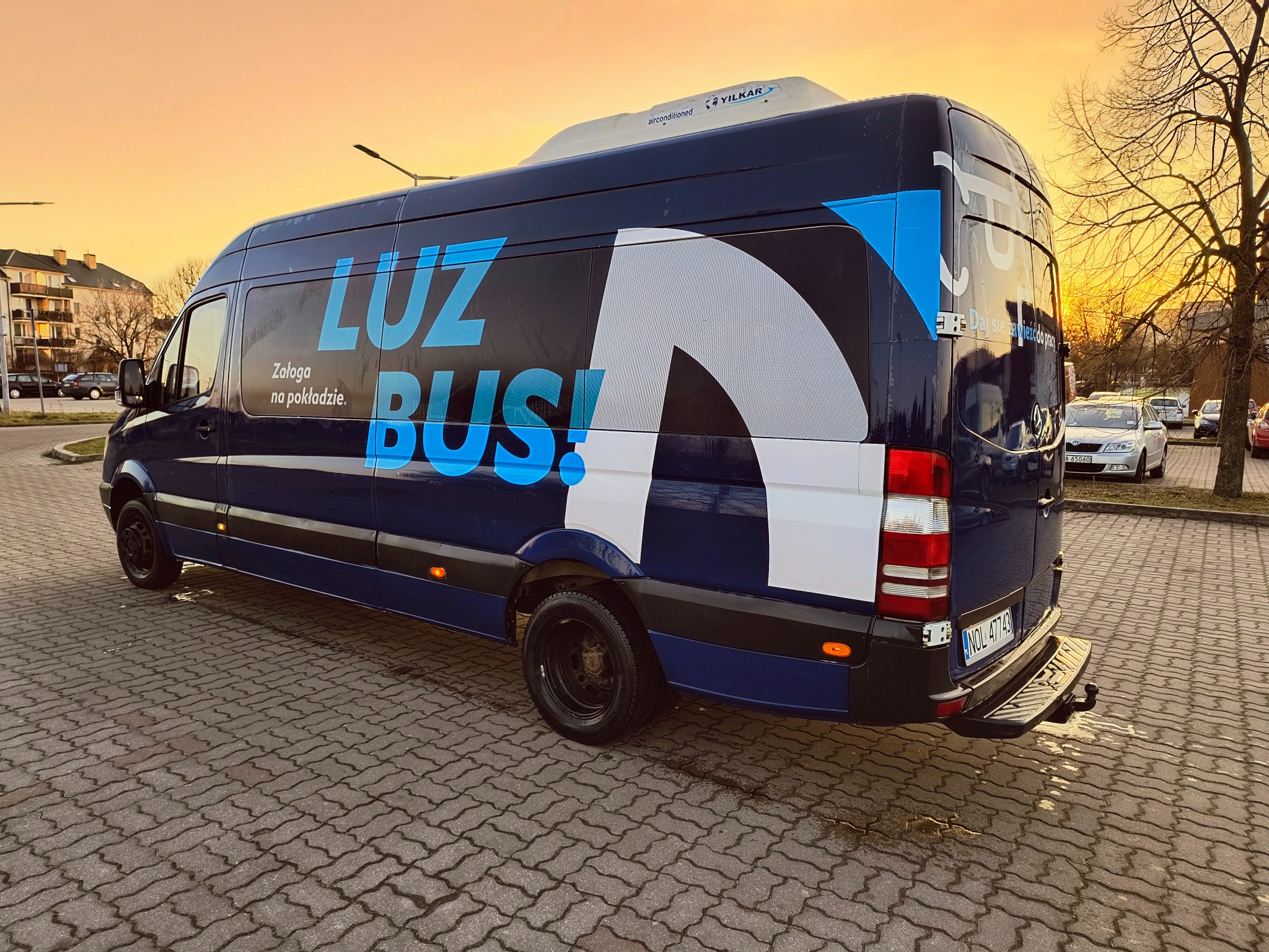 Niebieski bus z napisem 'LUZ BUS! Załoga na pokładzie' na tle parkingu i zachodzącego słońca.