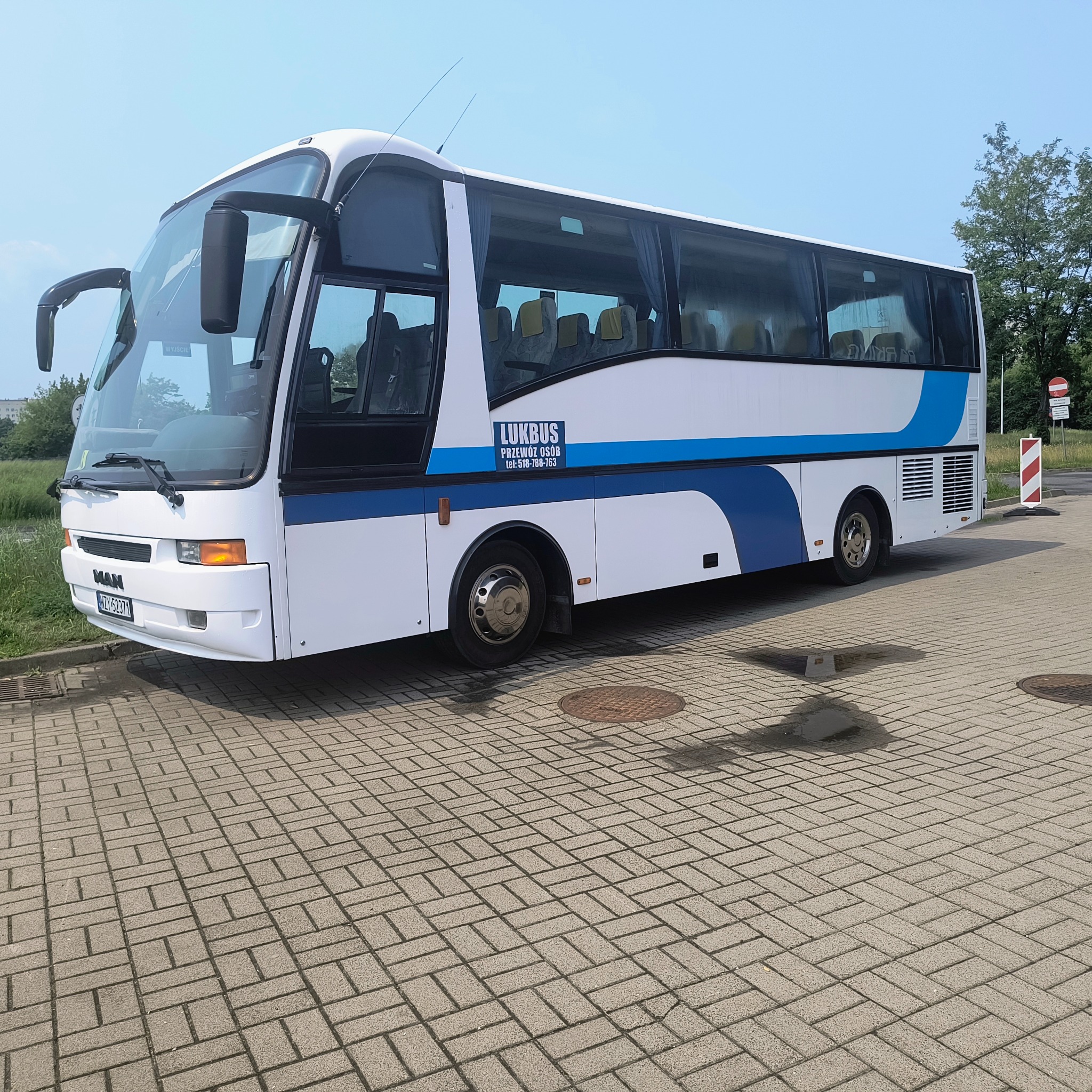 Biały autokar MAN z niebieskim pasem, zaparkowany na brukowanym placu. Widoczne logo 'LUKBUS' i numer telefonu. W tle zieleń i błękitne niebo.