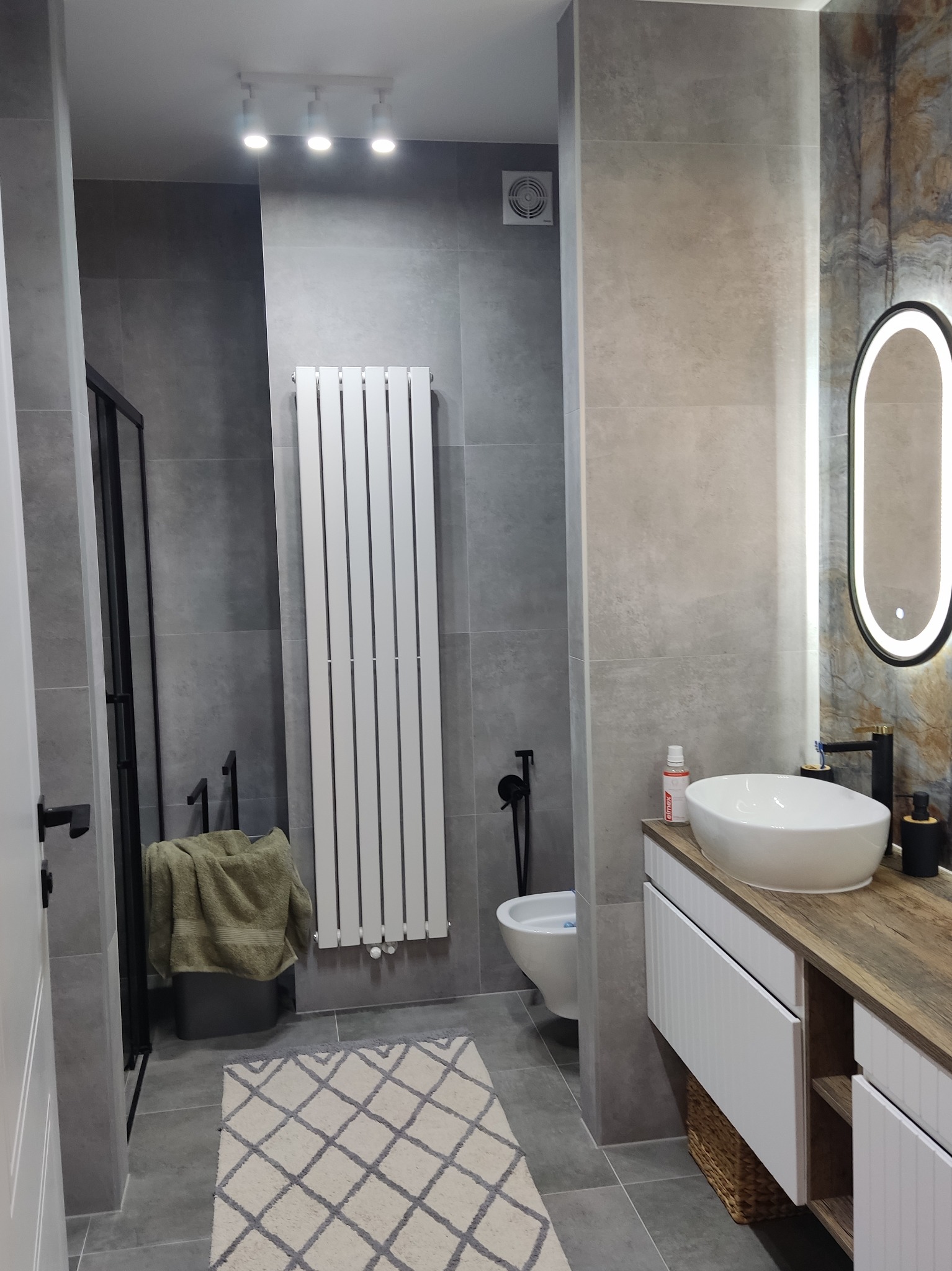 Nowoczesna łazienka w szarościach z białym grzejnikiem drabinkowym, owalnym lustrem z podświetleniem LED, białą umywalką nablatową na szafce z drewnianym blatem i czarną armaturą, widoczna bidetka...