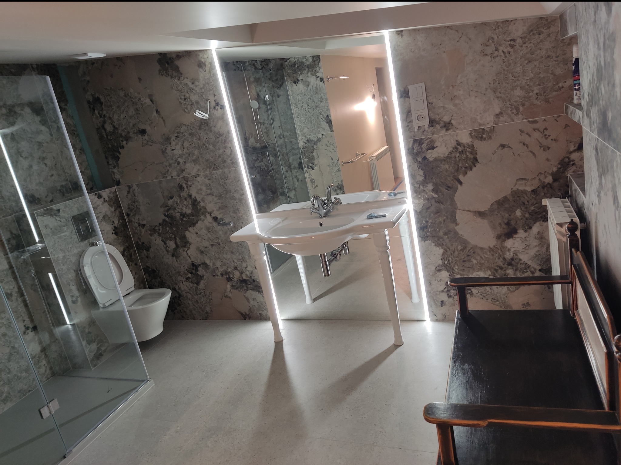 Nowoczesna łazienka z marmurowymi płytkami, kabiną prysznicową ze szkła, umywalką na ozdobnych nogach i czarną ławką.