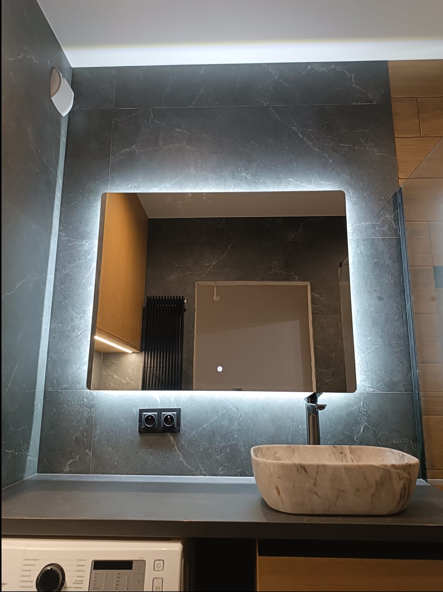 Nowoczesna łazienka z podświetlanym lustrem, umywalką z efektem marmuru i szarymi płytkami imitującymi kamień.
