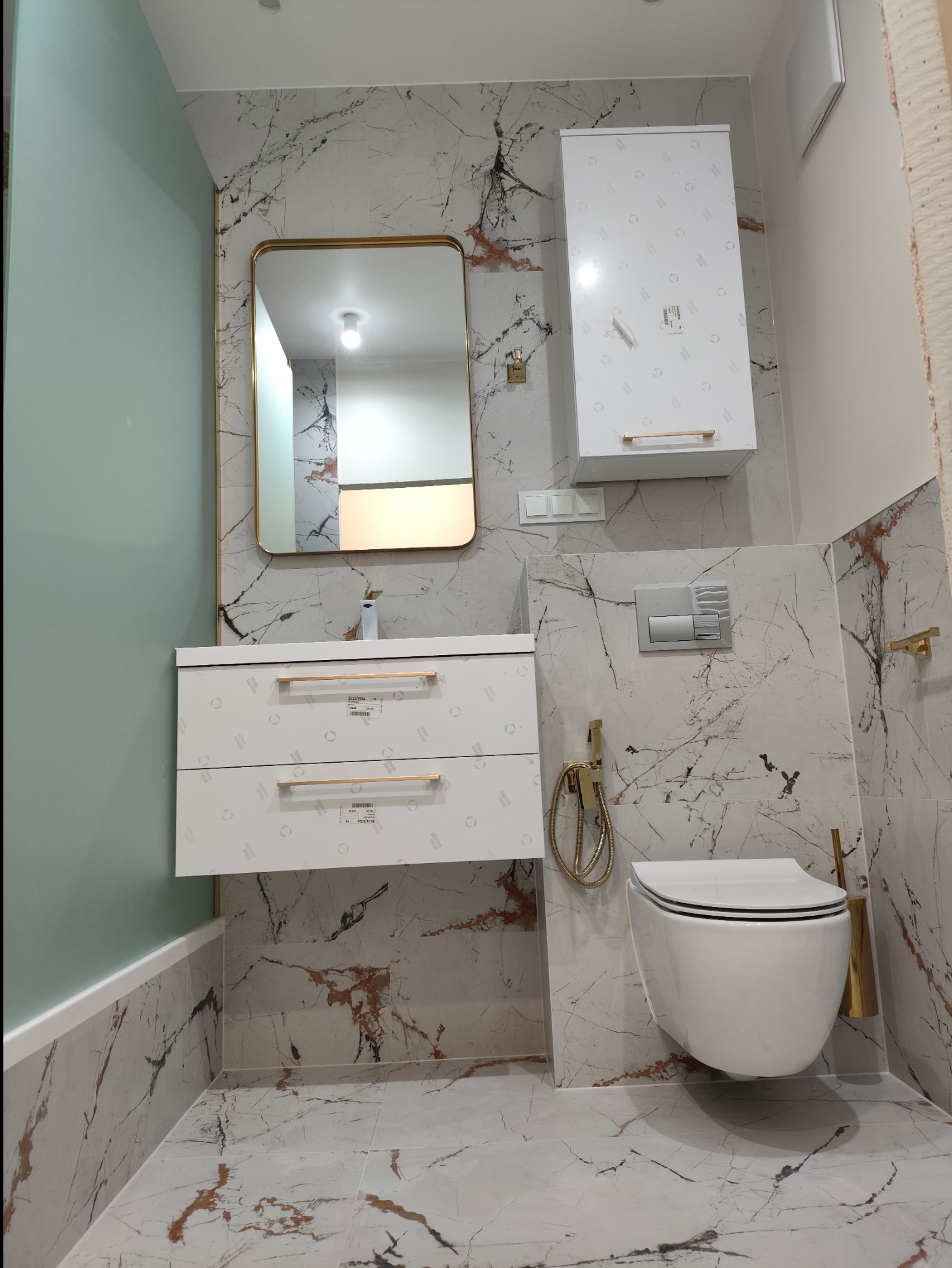 Nowoczesna łazienka z marmurowymi płytkami, wiszącą miską WC, szafką pod umywalkę z złotymi uchwytami i lustrem w złotej ramie.