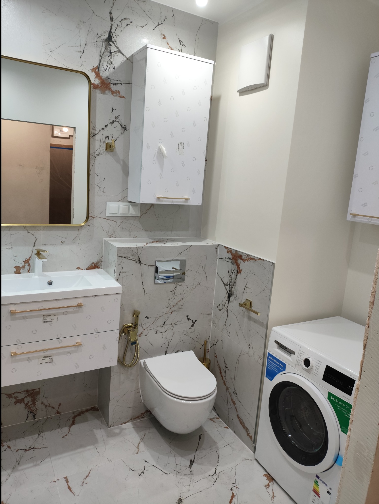 Nowoczesna łazienka z marmurowymi płytkami, wiszącą toaletą, białą umywalką z szafką, złotymi akcentami i pralką Bosch z serii 4.