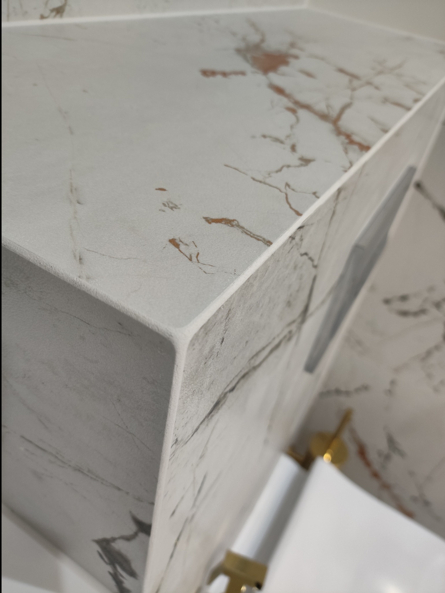 Narożnik blatu łazienkowego z białego marmuru z delikatnymi, złotymi żyłkami, wykończony z precyzją, widoczny z bliska, z fragmentem armatury w tle.