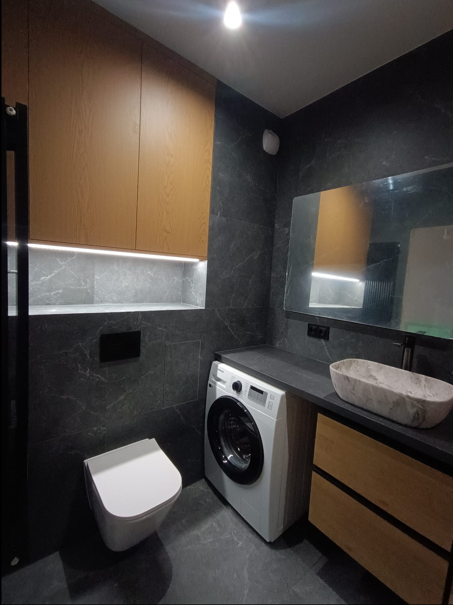 Nowoczesna łazienka z szarymi marmurkowymi płytkami, podświetlaną wnęką z półką, wiszącą toaletą, pralką i umywalką w kształcie łodzi na blacie z drewnianymi szafkami.