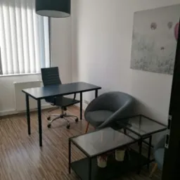 Wnętrze biura do wynajęcia: czarne biurko z krzesłem obrotowym, szare krzesło klubowe, stoliki kawowe z szklanym blatem, obraz z motywem balonów, rolety pionowe w oknie.