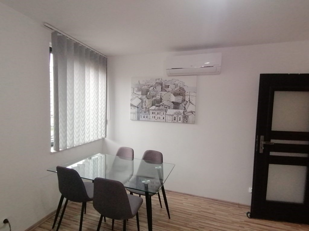 Jasne wnętrze z szklanym stołem, czterema krzesłami i obrazem z motywem miejskim na ścianie, klimatyzacja zamontowana pod sufitem, pionowe żaluzje w oknie.