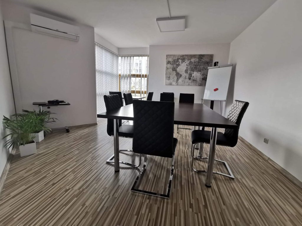 Sala konferencyjna z dużym stołem, czarnymi krzesłami z chromowanymi nogami, tablicą suchościeralną i mapą świata na ścianie. Rośliny doniczkowe w białych osłonkach w rogu pomieszczenia.