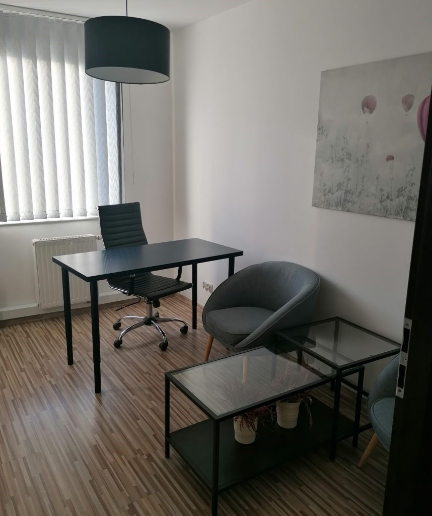 Wnętrze biura do wynajęcia: czarne biurko z krzesłem obrotowym, szare krzesło klubowe, stoliki kawowe z szklanym blatem, obraz z motywem balonów, rolety pionowe w oknie.
