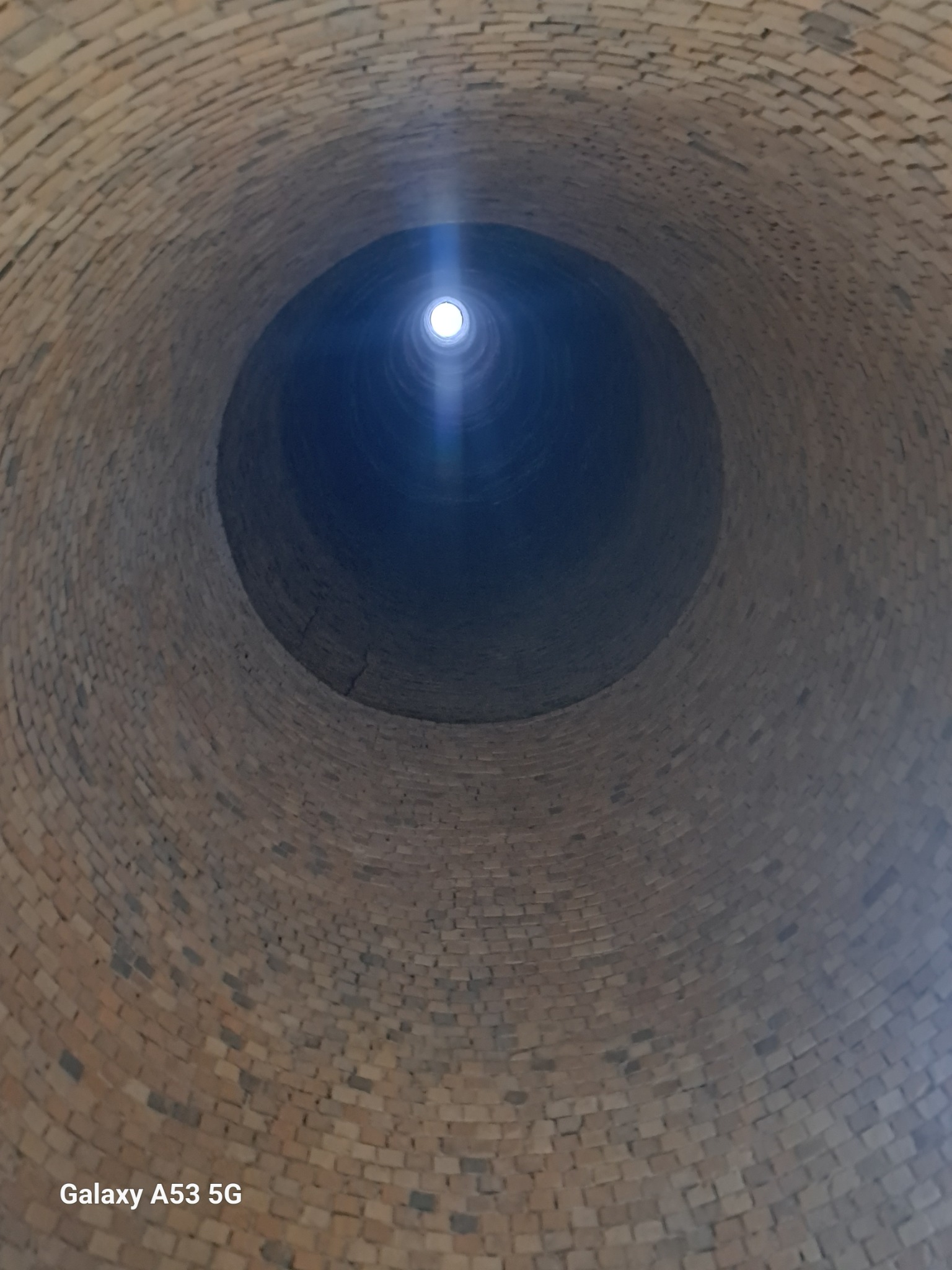 Wnętrze ceglanego komina przemysłowego widziane od dołu, z widocznym otworem w górnej części, przez który wpada jasne światło.