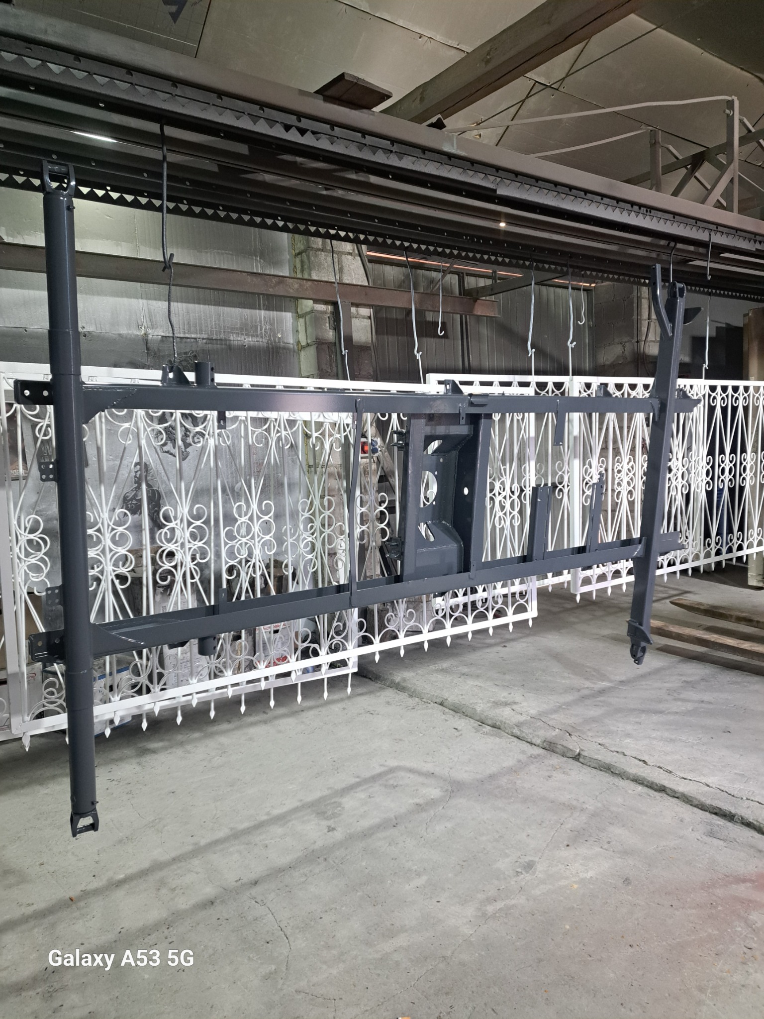 Metalowa konstrukcja bramy i inne elementy zawieszone na hakach w hali produkcyjnej, prawdopodobnie po procesie malowania lub czyszczenia, z widoczną białą, ozdobną bramą w tle.