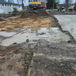 Żółty koparko-ładowarka JCB na placu budowy, w tle budynek gospodarczy i silosy, teren częściowo przygotowany, widoczne błoto i kałuże.