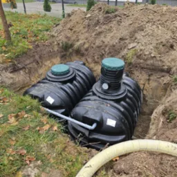 Dwa czarne zbiorniki z zielonymi pokrywami, połączone szarym rurociągiem, umieszczone w wykopie ziemnym na działce z trawą i opadłymi liśćmi, widoczny fragment izolowanej rury.