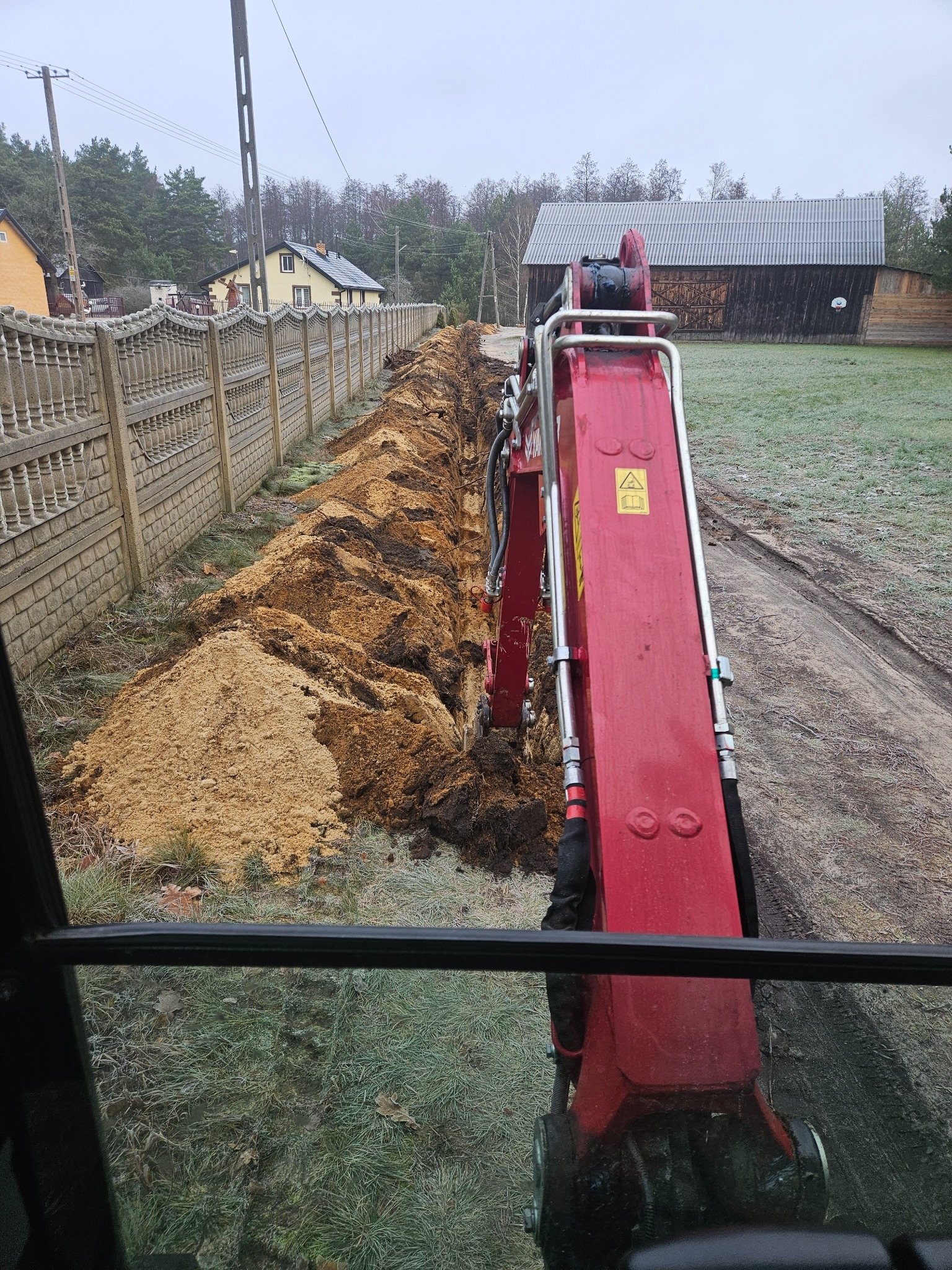 Wykopany rów wzdłuż ozdobnego betonowego ogrodzenia, widok z kabiny czerwonej minikoparki na tle zabudowań wiejskich i zroszonej trawy.