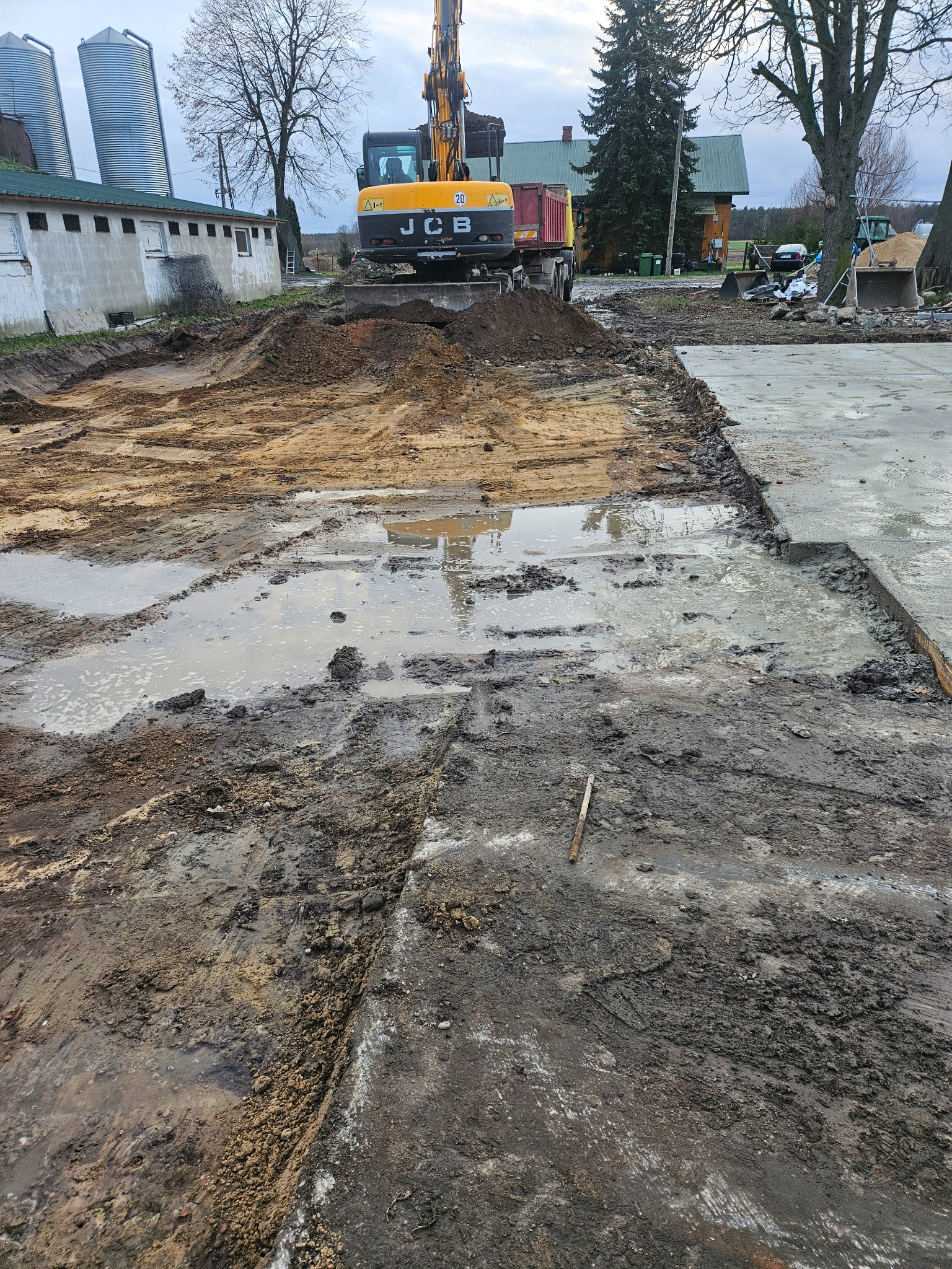 Żółty koparko-ładowarka JCB na placu budowy, w tle budynek gospodarczy i silosy, teren częściowo przygotowany, widoczne błoto i kałuże.