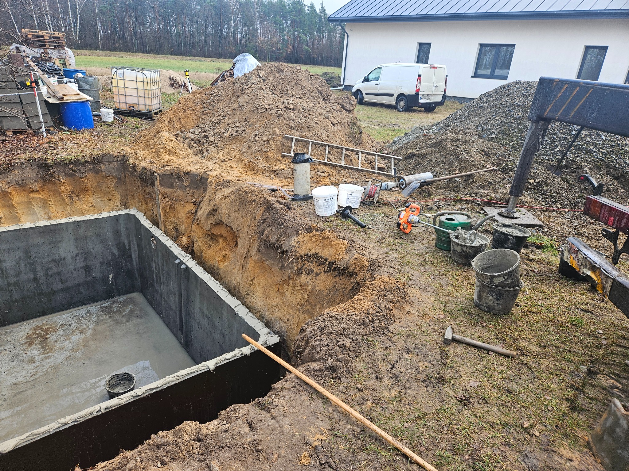 Betonowy zbiornik z wylanym dnem w wykopie, widoczne narzędzia budowlane, hałda ziemi i budynek w tle.