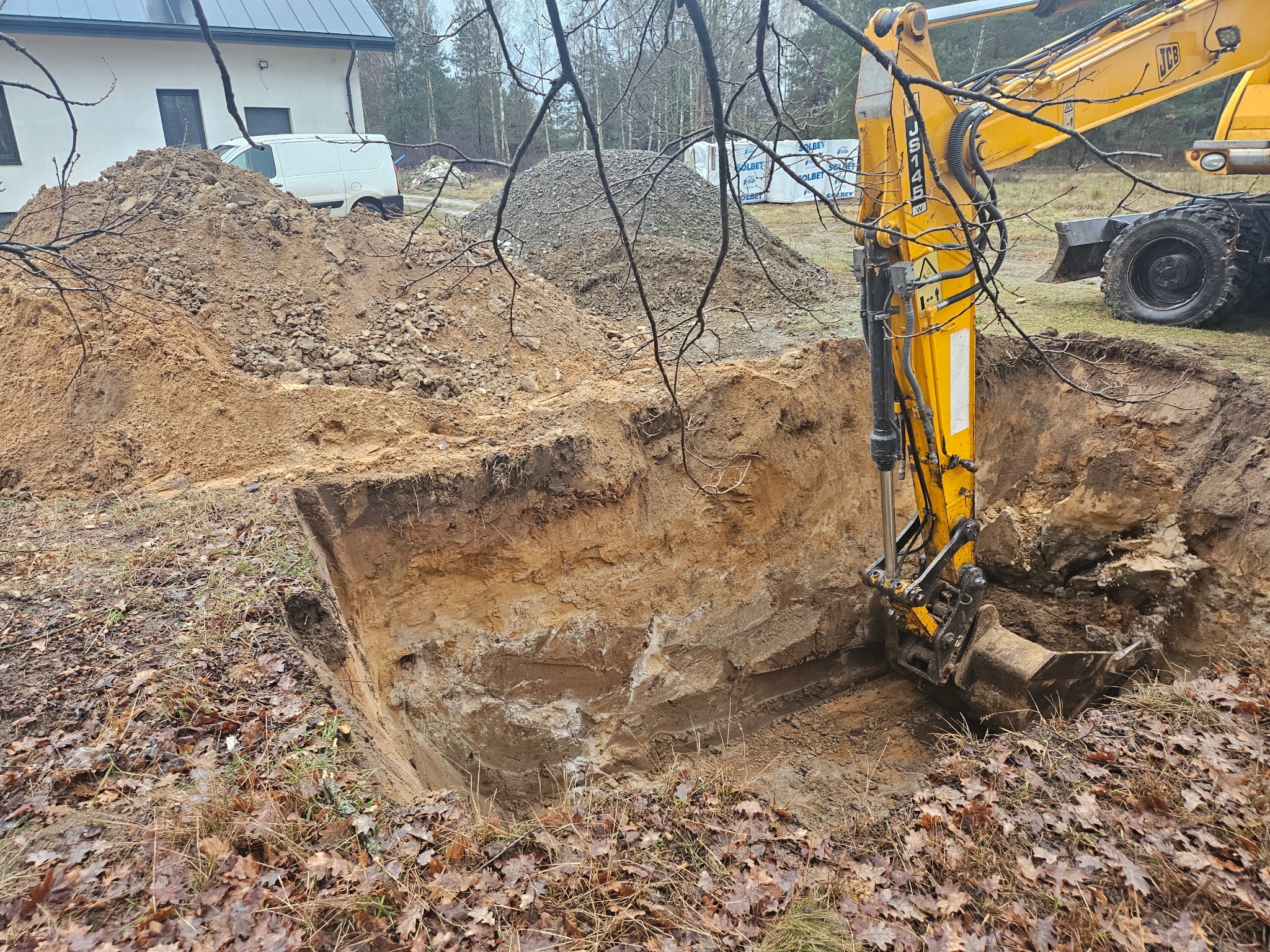 Żółta koparka JCB w trakcie pracy przy wykopie o nierównych ścianach, obok hałda ziemi i budynek z białymi ścianami, w tle drzewa.