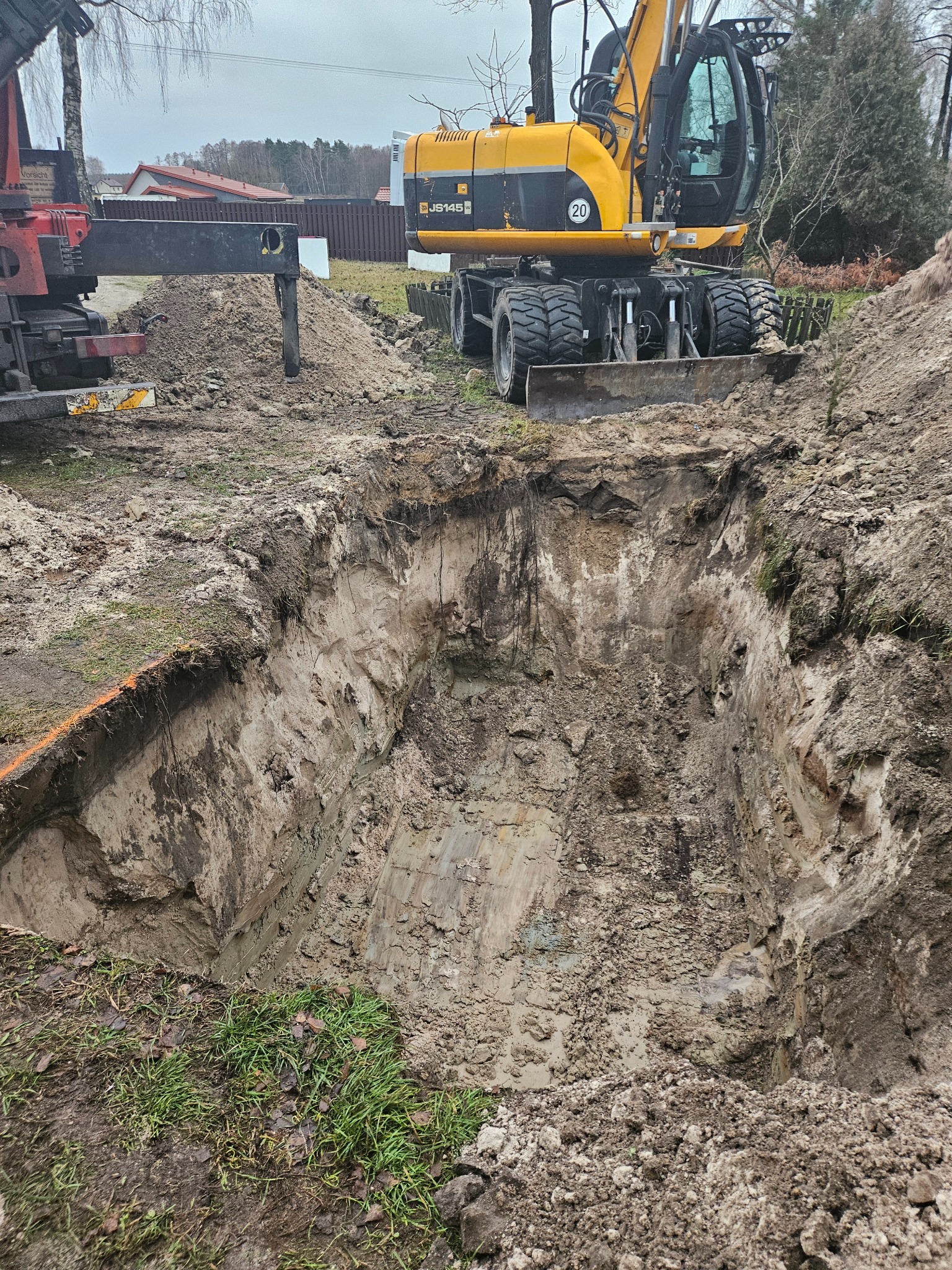 Głęboki wykop w ziemi, z żółtą koparką JCB na kołach w tle, obok widoczny fragment żurawia samochodowego, pomarańczowa linia wyznacza krawędź wykopu.