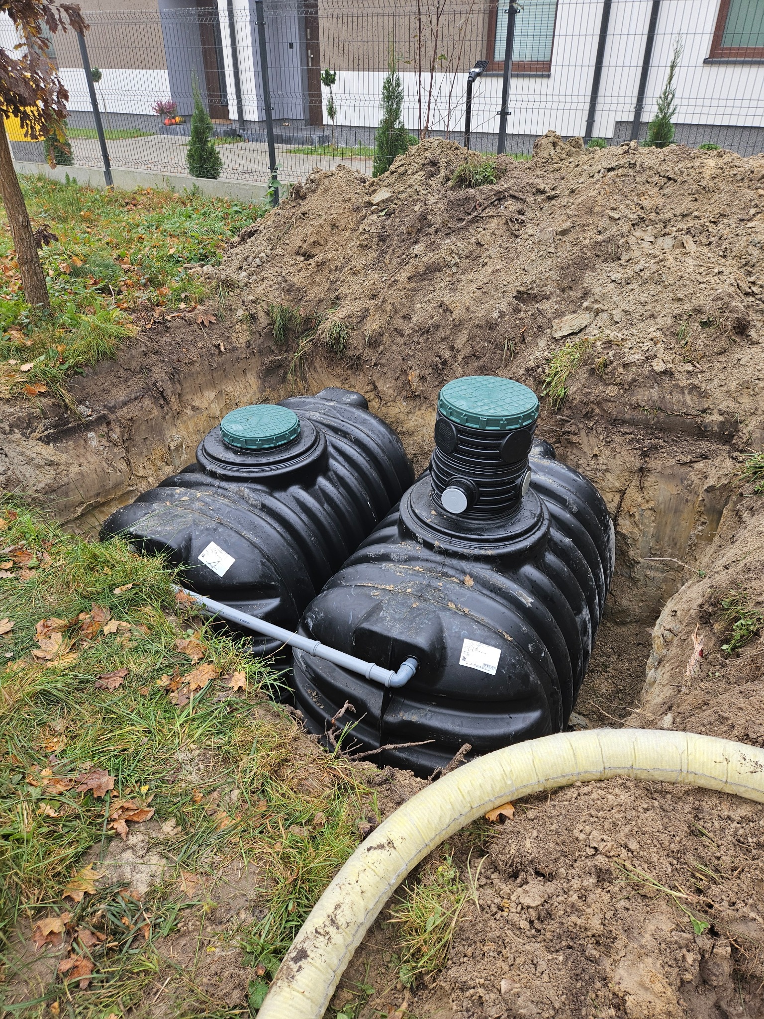 Dwa czarne zbiorniki z zielonymi pokrywami, połączone szarym rurociągiem, umieszczone w wykopie ziemnym na działce z trawą i opadłymi liśćmi, widoczny fragment izolowanej rury.