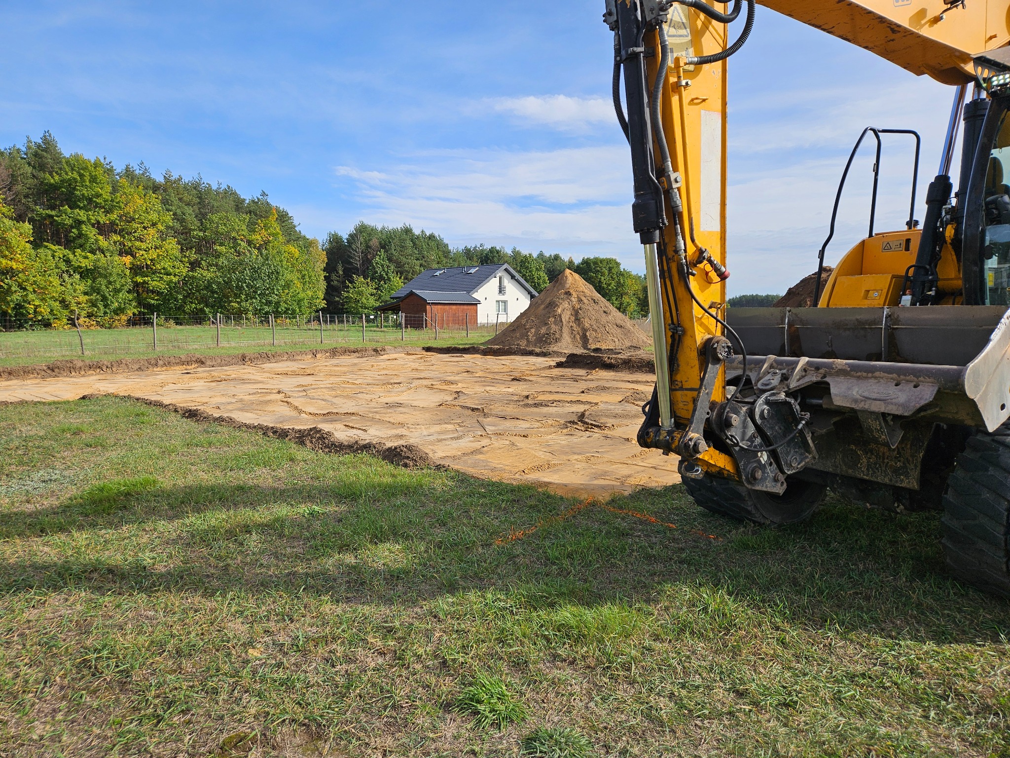 Żółta koparka na działce z przygotowanym terenem pod budowę, widoczny wyznaczony obrys fundamentów, w tle dom jednorodzinny i sterta ziemi.