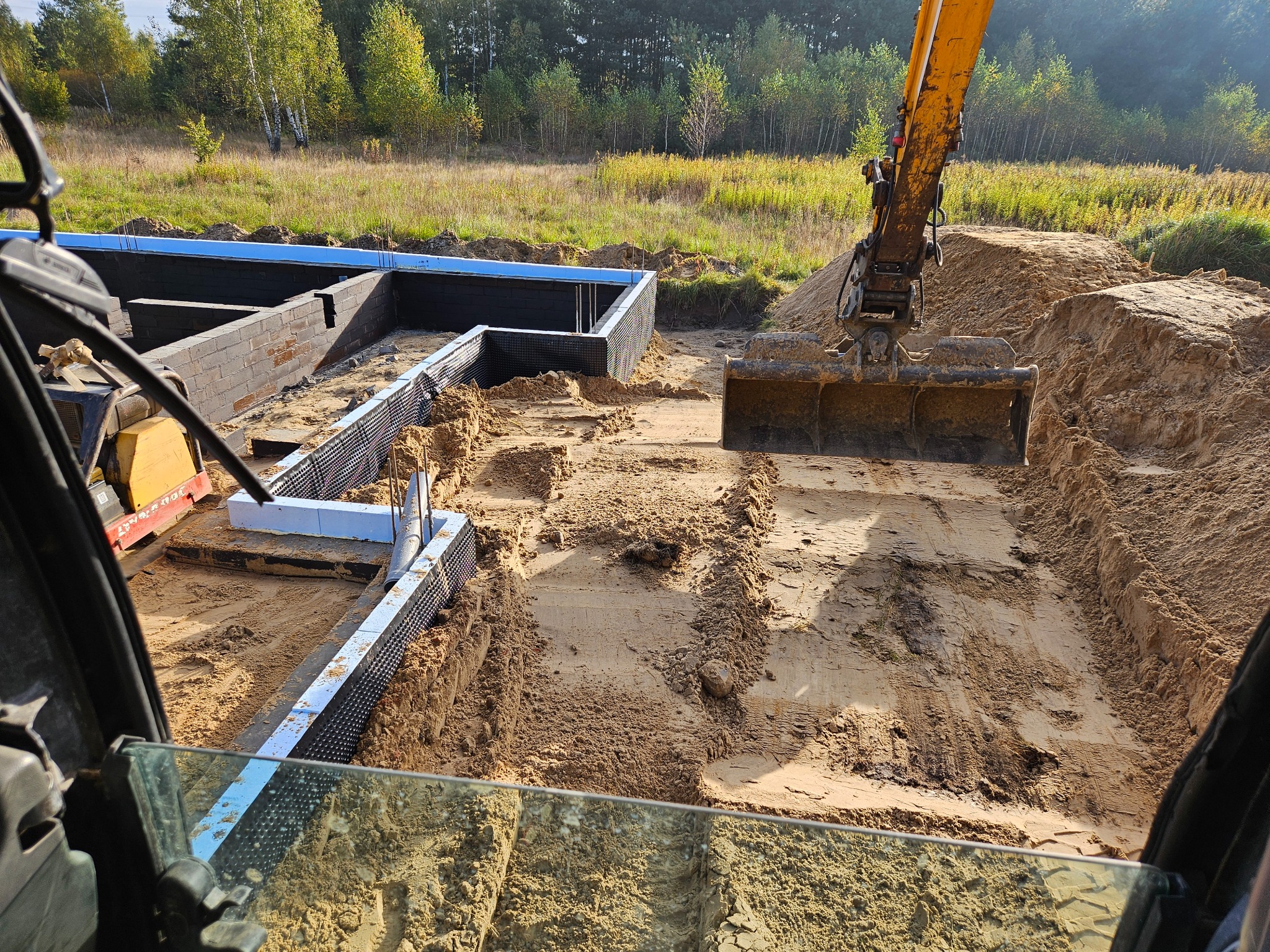 Żółta koparka wyrównuje dno wykopu pod fundamenty budynku, widok z kabiny operatora na częściowo ukończone ściany fundamentowe z izolacją pionową i niebieskim obrzeżem, w tle drzewa.