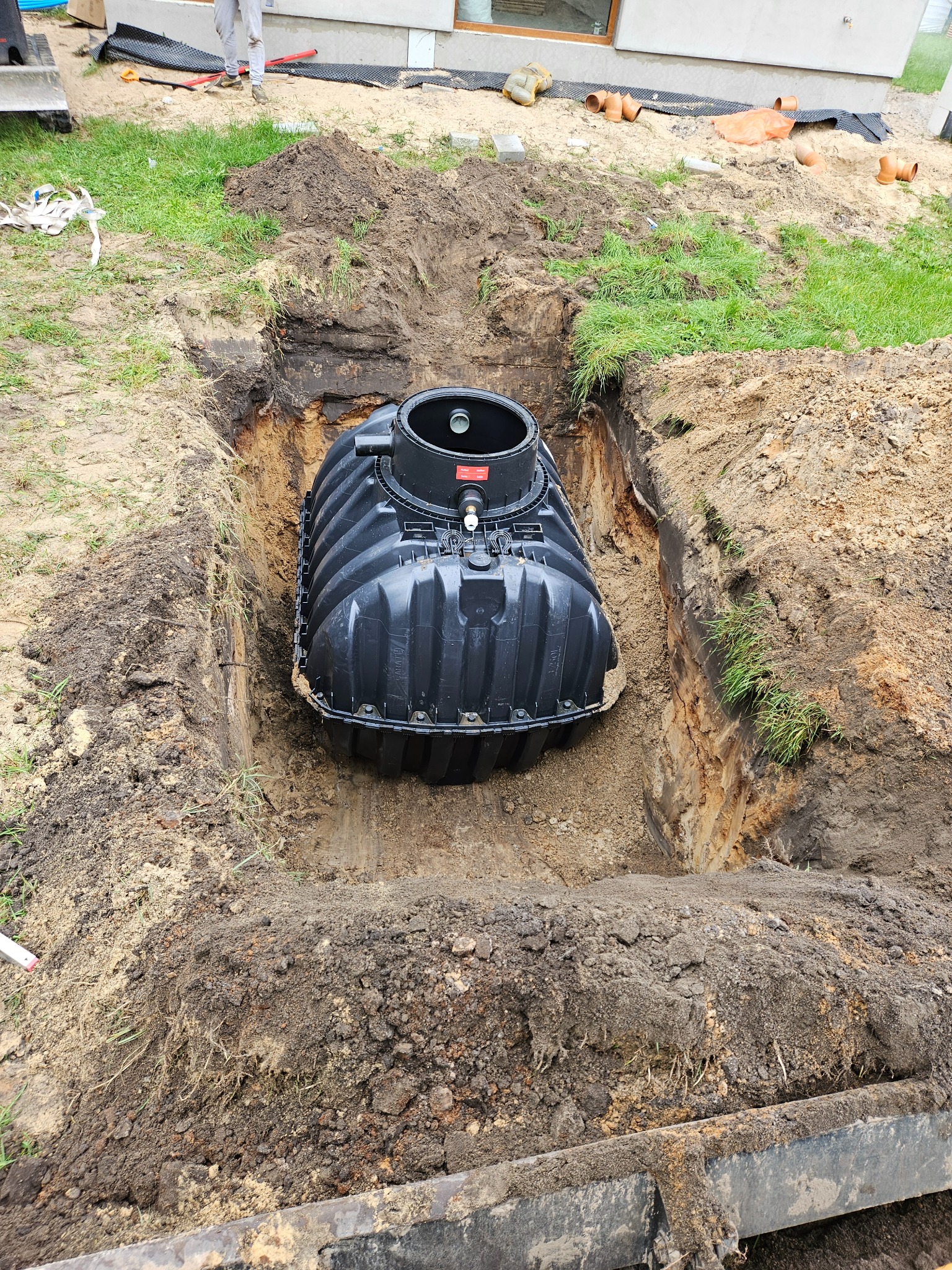 Czarny zbiornik z tworzywa sztucznego umieszczony w wykopie ziemnym, widoczne ślady prac ziemnych i trawa na powierzchni gruntu, elementy instalacji sanitarnej w tle.
