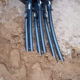 Czarna studzienka z podłączonymi rurami w piasku, widoczne ślady butów. Instalacja pompy ciepła w ziemi, krok w procesie instalacyjnym. Detal przyłącza.