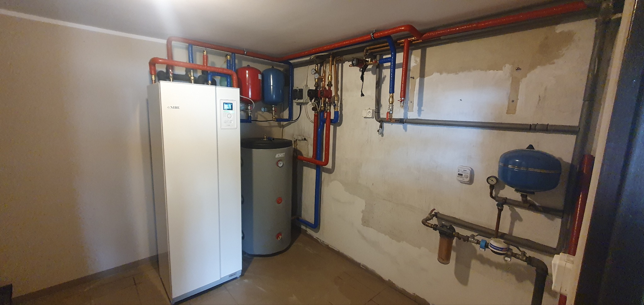 Kompletna instalacja grzewcza z pompą ciepła Nibe, zbiornikami buforowymi i rurami w kolorach czerwonym i niebieskim. Widoczne liczniki i osprzęt.