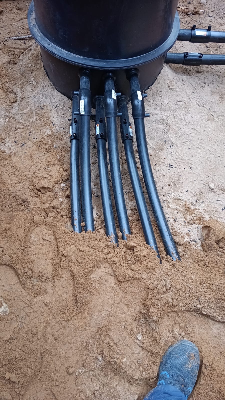 Czarna studzienka z podłączonymi rurami w piasku, widoczne ślady butów. Instalacja pompy ciepła w ziemi, krok w procesie instalacyjnym. Detal przyłącza.
