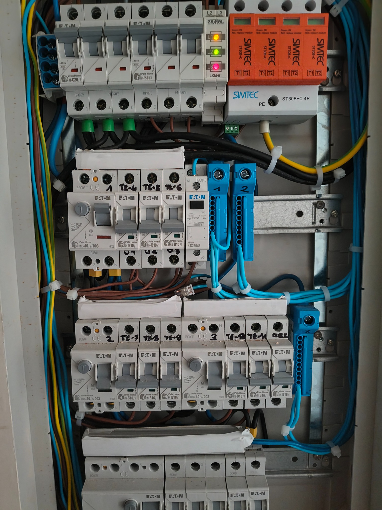 Rozdzielnica elektryczna z zainstalowanymi bezpiecznikami, modułami przeciwprzepięciowymi Simtec i okablowaniem w kolorach niebieskim, żółtym i brązowym.