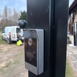 PJserwis Mateusz Jagoda - Zamontowany wideodomofon marki EZVIZ na czarnym słupku bramy, z widocznym pojazdem serwisowym w tle i elementami instalacji.