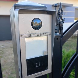 PJserwis Mateusz Jagoda - Zewnętrzny panel wideodomofonu Hikvision zamontowany na czarnym, kutym ogrodzeniu, z widocznym odbiciem otoczenia w obiektywie kamery.
