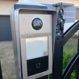 Zewnętrzny panel wideodomofonu Hikvision zamontowany na czarnym, kutym ogrodzeniu, z widocznym odbiciem otoczenia w obiektywie kamery.