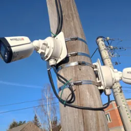 Dwie kamery monitoringu marki Internec zamontowane na drewnianym słupie za pomocą metalowych obejm, widoczne przewody i fragment budynku w tle, błękitne niebo.