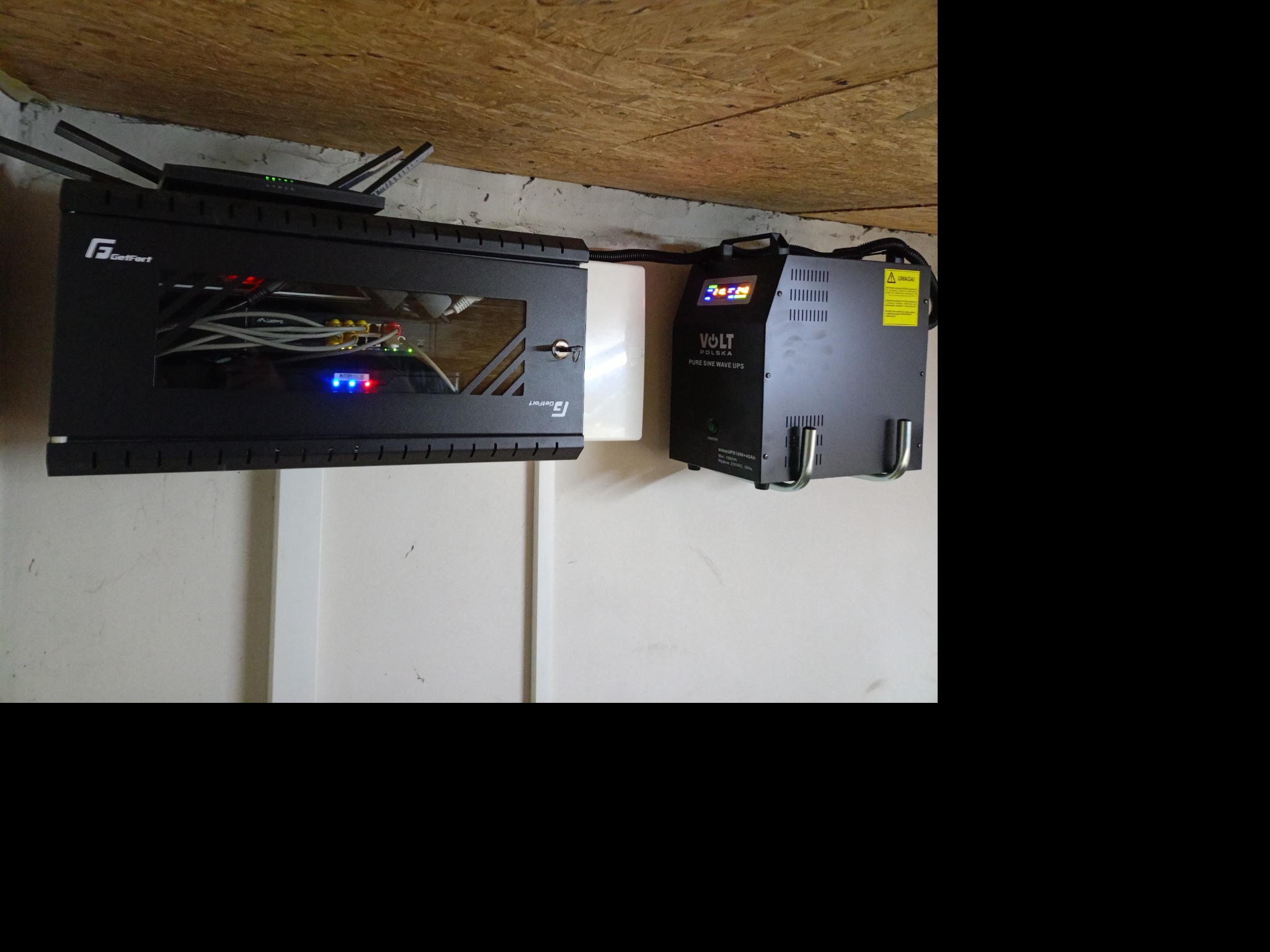 Szafa rack GetFort z routerem i okablowaniem sieciowym, obok zasilacz awaryjny UPS Volt Polska z wyświetlaczem, zamontowane na ścianie pod sufitem.
