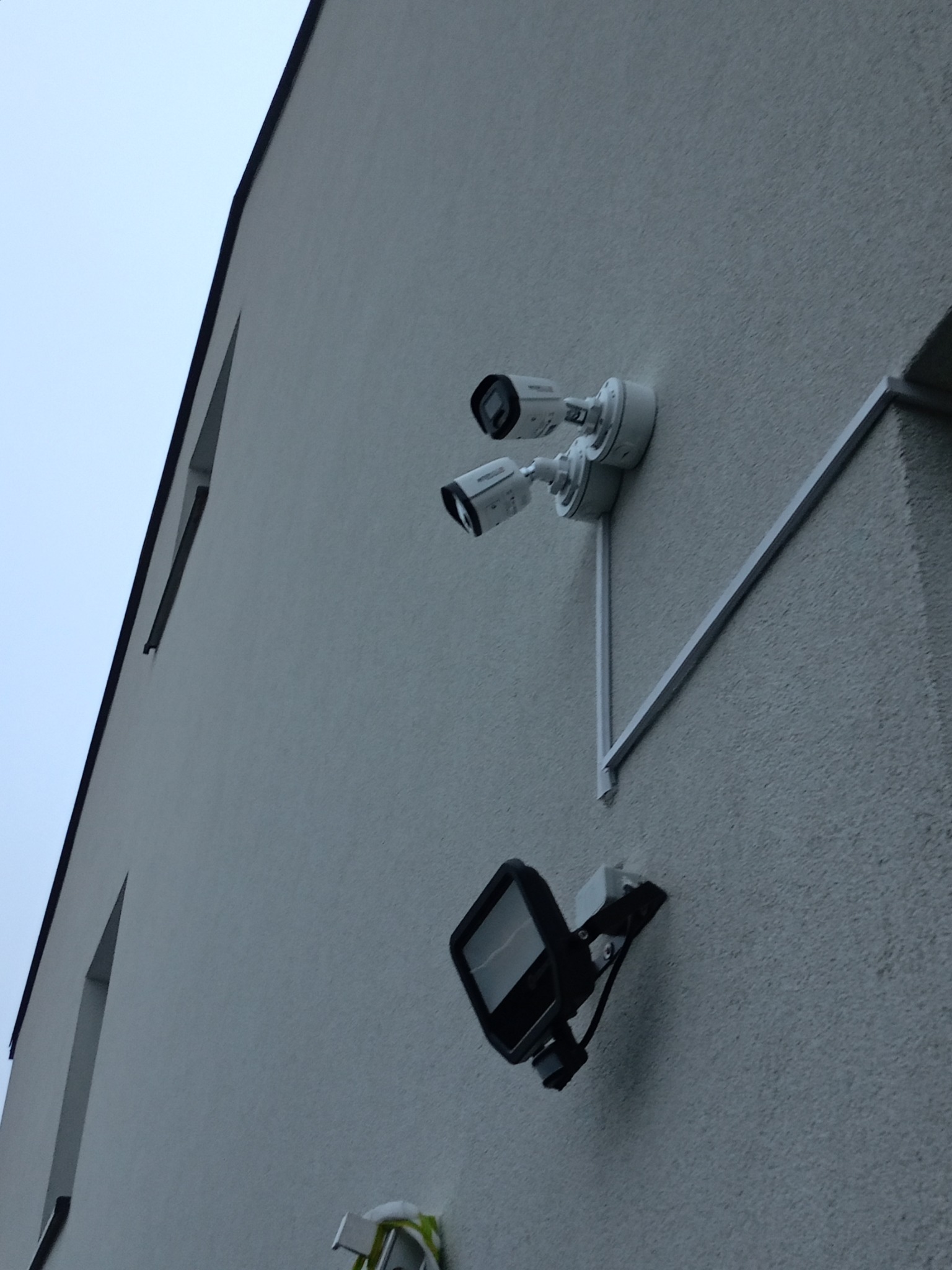 Dwie kamery monitoringu przymocowane do elewacji budynku, poniżej lampa oświetleniowa, widok z dołu na szarą ścianę i pochmurne niebo.