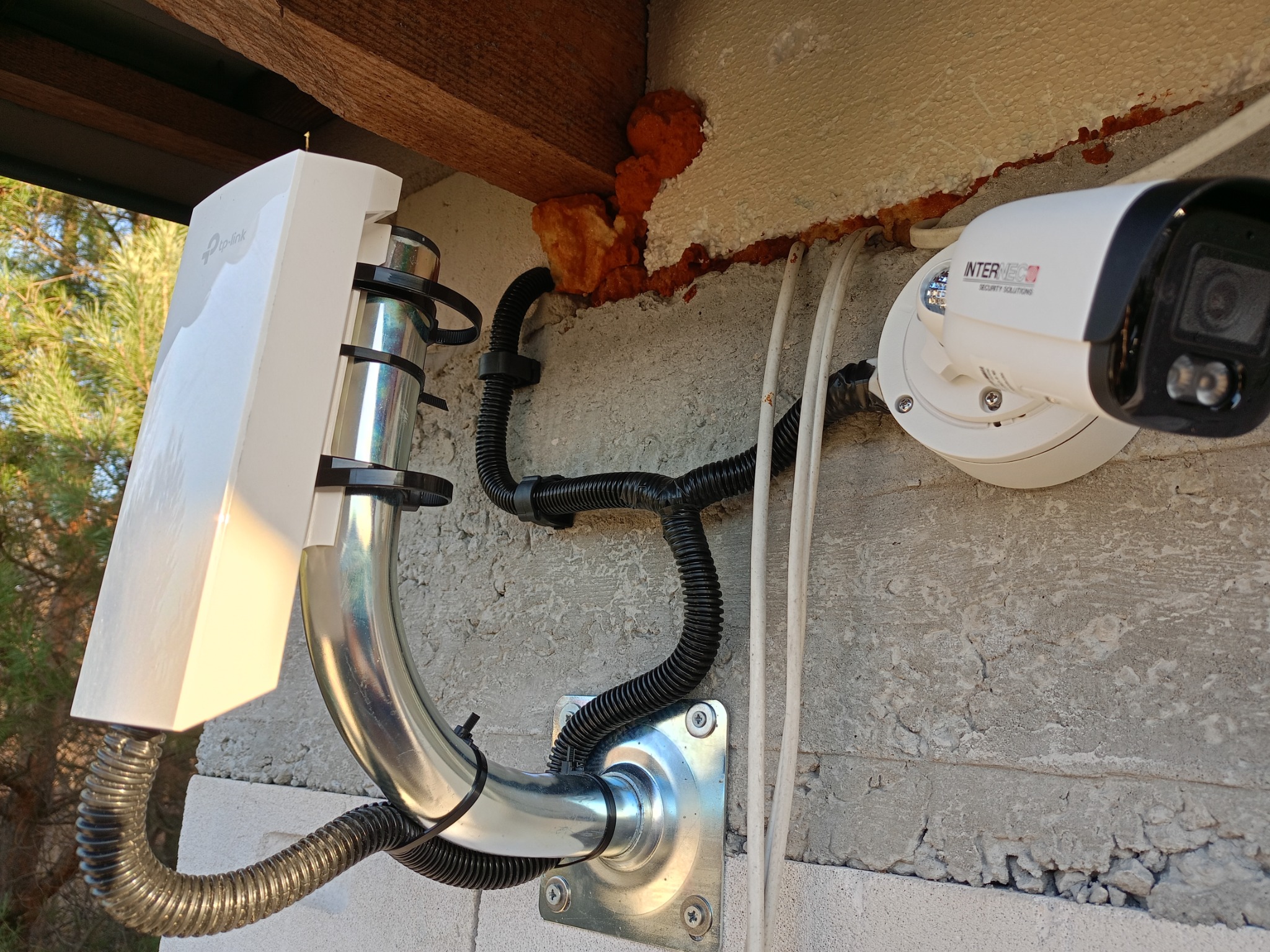Zewnętrzna instalacja kamery monitoringu Intermec Security Solutions z widocznym routerem TP-Link, przymocowana do ściany budynku z użyciem metalowej rury i peszli ochronnych.