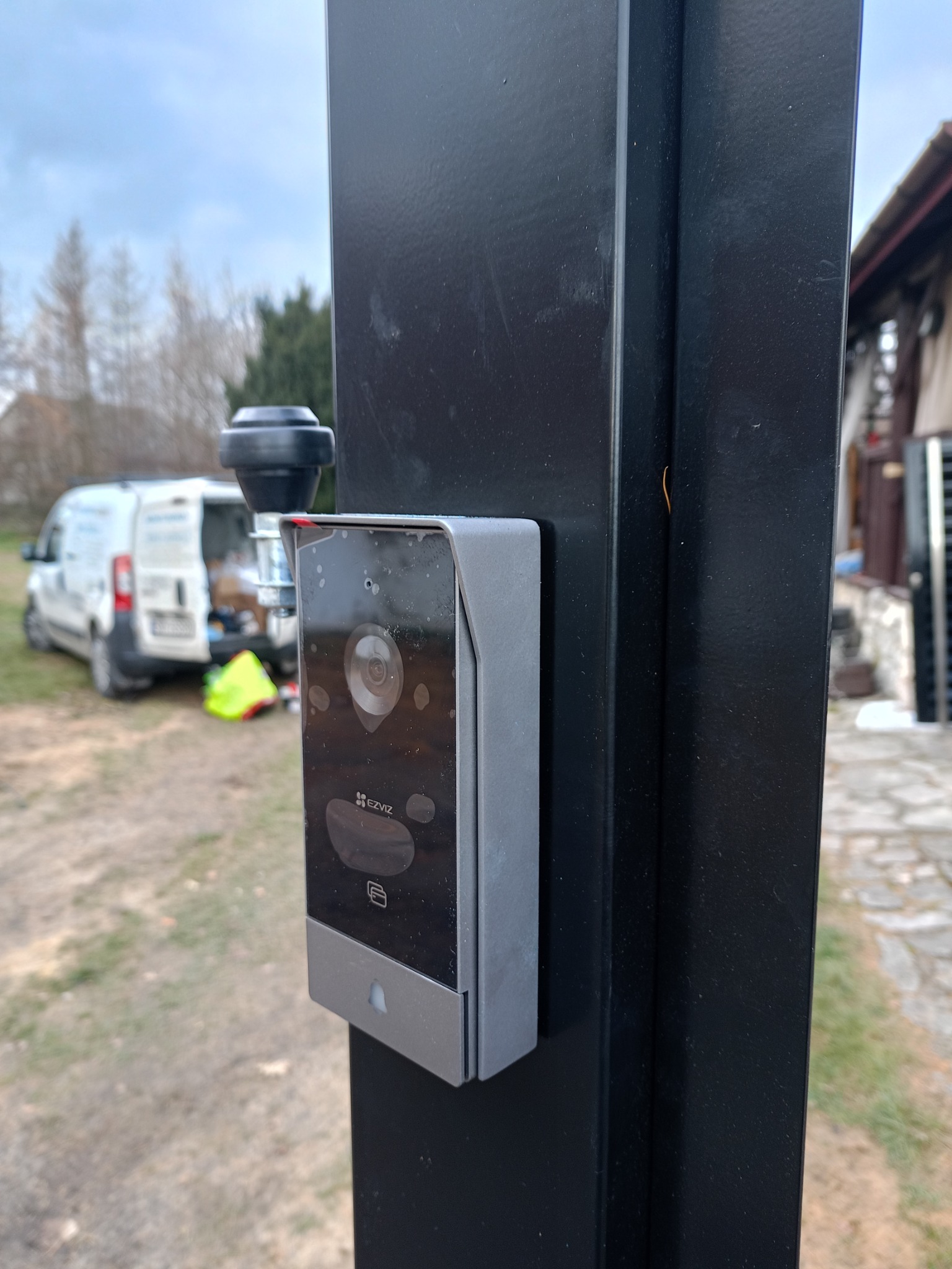 Zamontowany wideodomofon marki EZVIZ na czarnym słupku bramy, z widocznym pojazdem serwisowym w tle i elementami instalacji.