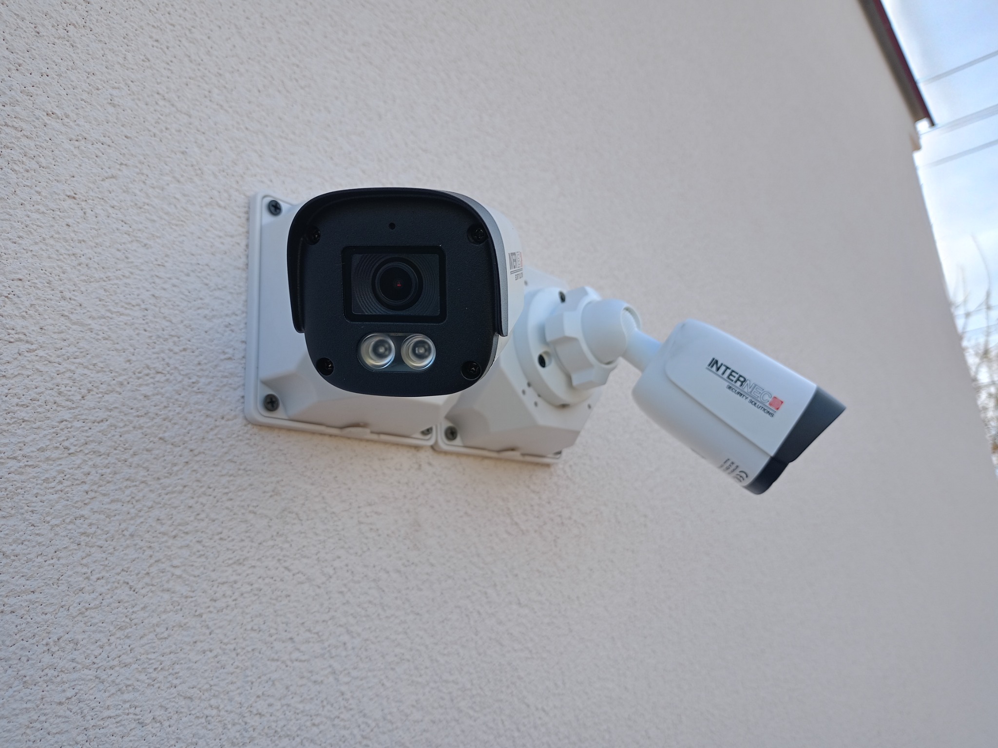 Zewnętrzna kamera monitoringu z dwoma diodami IR, zamontowana na jasnej, teksturowanej ścianie budynku, widoczny fragment elewacji i logo producenta.