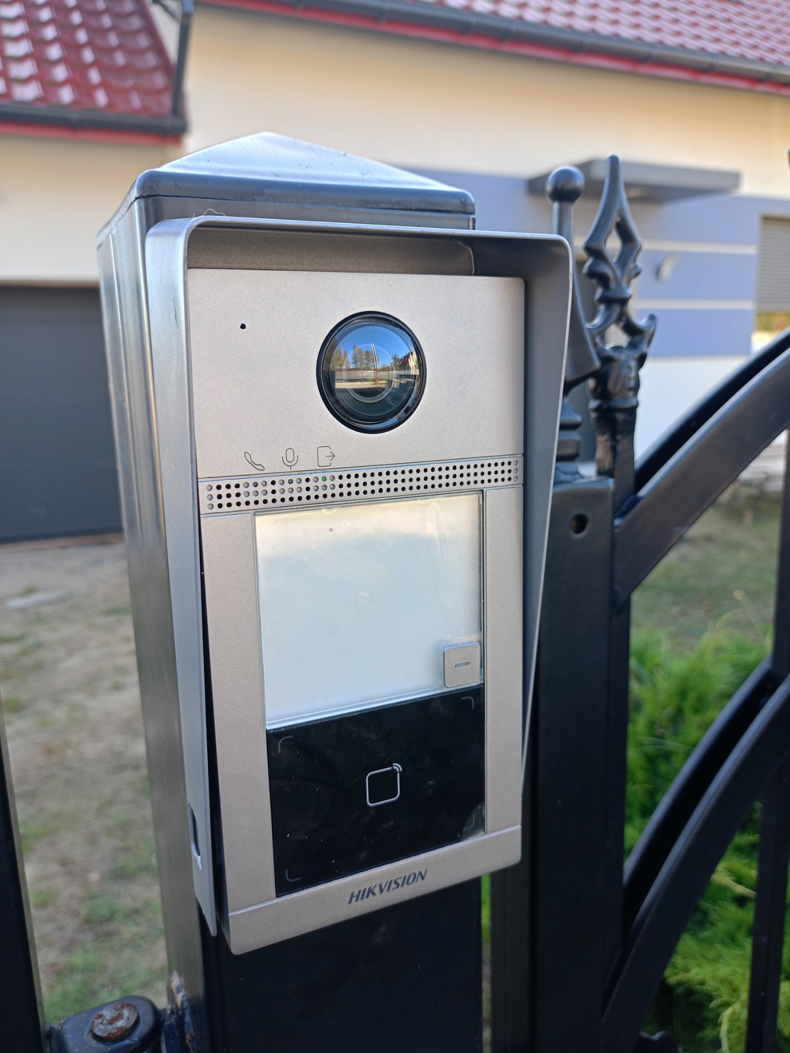 Zewnętrzny panel wideodomofonu Hikvision zamontowany na czarnym, kutym ogrodzeniu, z widocznym odbiciem otoczenia w obiektywie kamery.