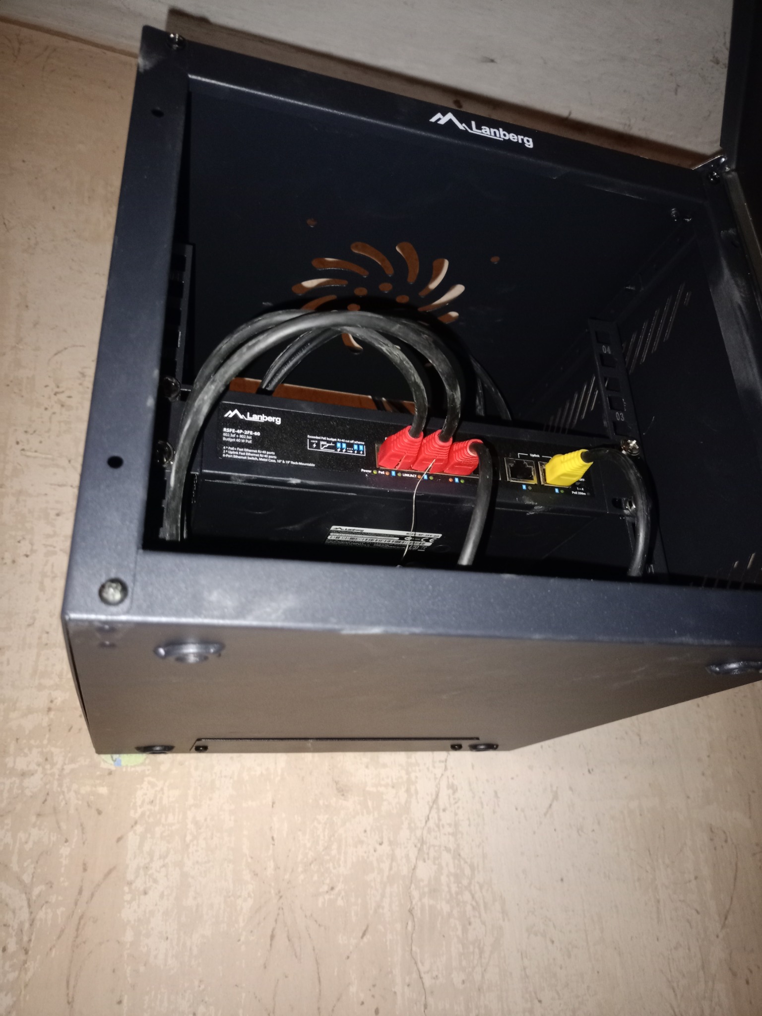 Czarny switch Lanberg w szafce rack, z widocznymi kablami Ethernet z czerwonymi i żółtymi końcówkami, zamontowany na ścianie z wzorzystą tapetą.