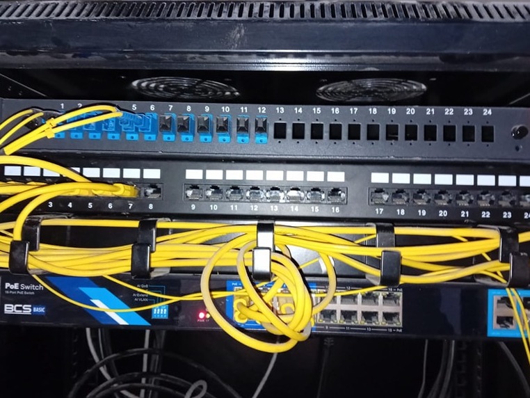 Panel krosowniczy z podłączonymi żółtymi kablami sieciowymi, widoczny switch PoE BCS Basic w szafie rack.