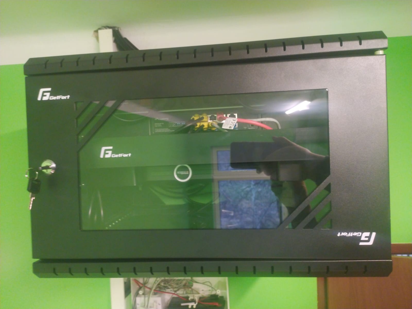 Zamykana na klucz szafka rack wisząca na zielonej ścianie, z widocznym okablowaniem sieciowym i elementami montażowymi wewnątrz, odbicie osoby w szybie.