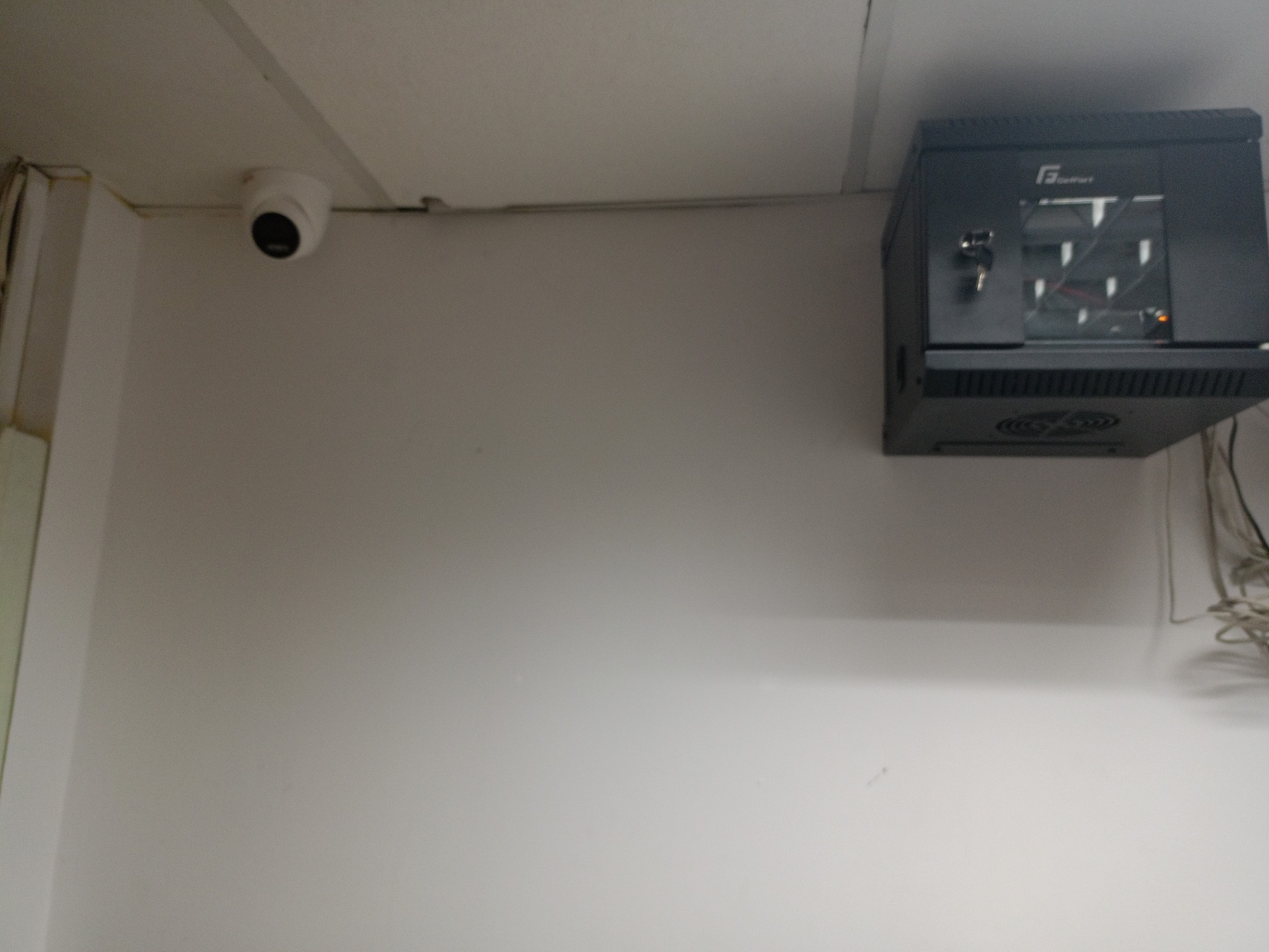 Kamera kopułkowa monitoringu zamontowana pod sufitem obok szafy rack z kluczykiem w zamku, widoczne kable.