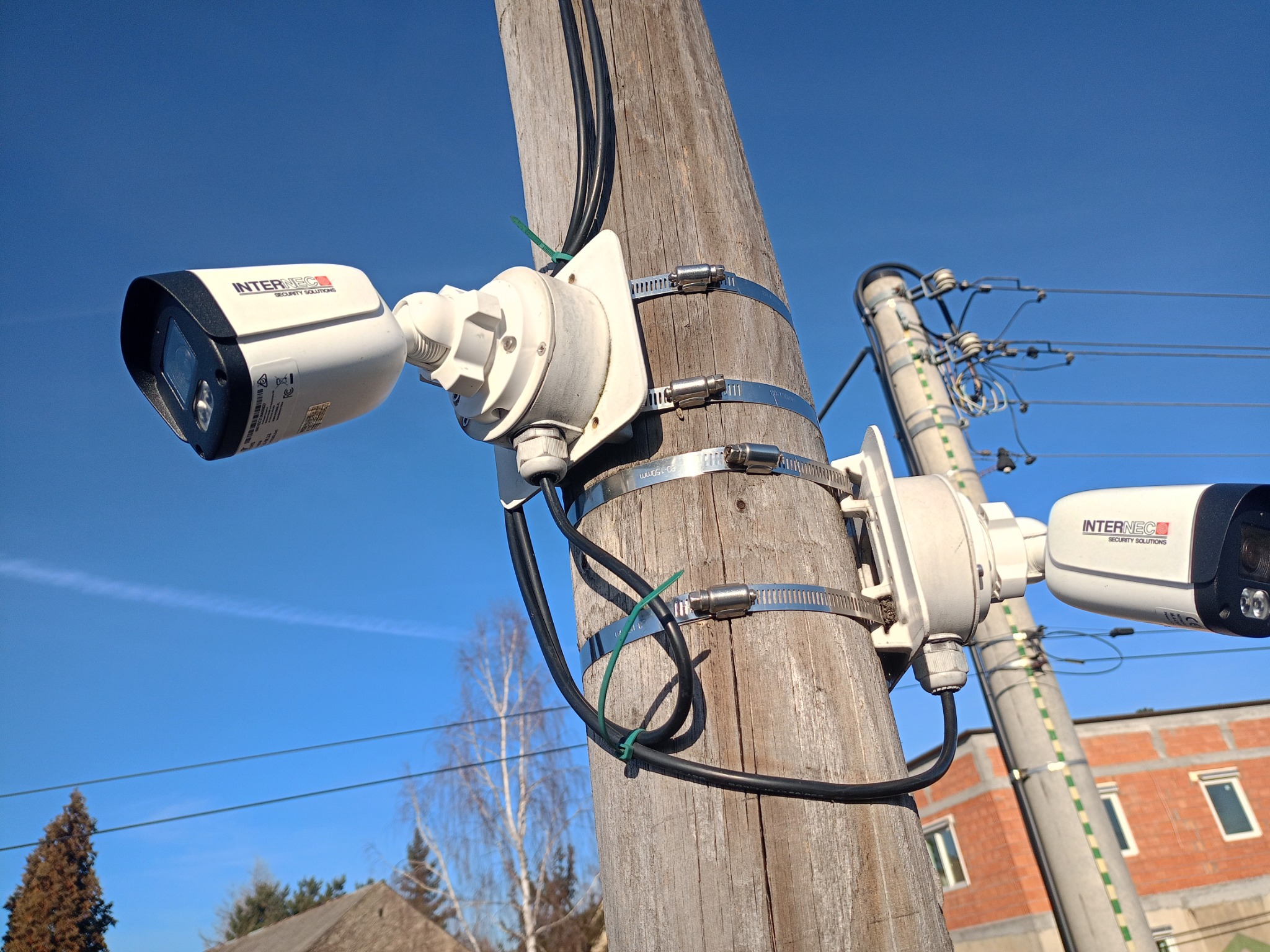 Dwie kamery monitoringu marki Internec zamontowane na drewnianym słupie za pomocą metalowych obejm, widoczne przewody i fragment budynku w tle, błękitne niebo.